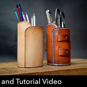 May include: Two cylindrical leather pencil holders with visible white stitching. The left holder is light tan and contains coloured pencils. The right holder is dark brown, features two horizontal straps with metal rivets, and holds scissors.