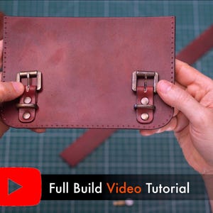 May include: A reddish-brown leather satchel with two brass buckles. The satchel is held by hands, with a strap visible in the background. The image includes a YouTube logo and the text "Full Build Video Tutorial".
