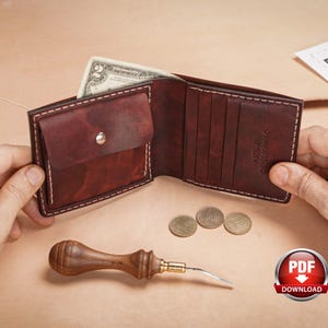 May include: A dark red leather bifold wallet with white stitching, featuring a coin pocket with a silver snap button, multiple card slots, and a debossed logo. A two-dollar bill is tucked inside, and a wooden handle tool lies nearby.