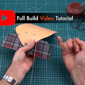 May include: A close-up of a person's hands sewing together a leather and plaid fabric wallet. The leather is a light brown color and the plaid fabric is red, green, and white. The person is using a needle and thread to sew the two pieces together. The background is a green cutting mat.