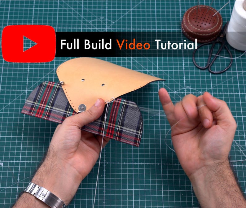 May include: A close-up of a person's hands sewing together a leather and plaid fabric wallet. The leather is a light brown color and the plaid fabric is red, green, and white. The person is using a needle and thread to sew the two pieces together. The background is a green cutting mat.