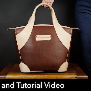 May include: A brown leather handbag featuring light beige leather accents on the handles, corners, and a central rectangular patch. The patch displays the text "DEELPUNTO". The bag has a zipper closure and white stitching along the edges.