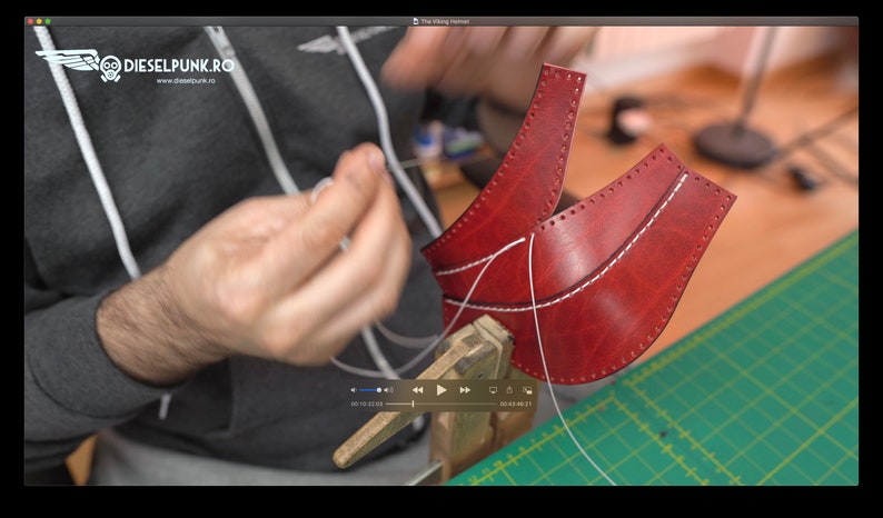 May include: A person is sewing two pieces of red leather together with white thread. The leather is clamped in a vise. The person is wearing a gray hoodie with the text "DIESELPUNK.RO" and the website address "www.dieselpunk.ro".