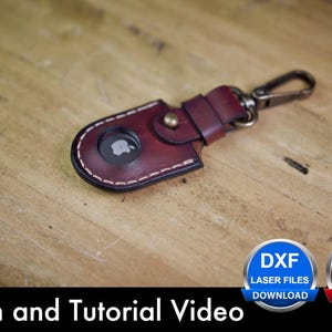 Op de afbeelding: Een donkerrode lederen sleutelhanger voor een Apple AirTag met witte stiksels, een messingkleurige drukknoop en een metalen karabijnhaak. De afbeelding bevat ronde iconen met de tekst DXF LASER FILES DOWNLOAD en PDF DOWNLOAD, en de tekst Pattern and Tutorial Video.