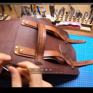 May include: A brown leather backpack with a top handle and adjustable straps. The backpack is being stitched together by hand.