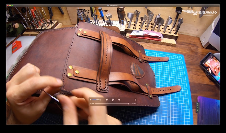 May include: A brown leather backpack with a top handle and adjustable straps. The backpack is being stitched together by hand.