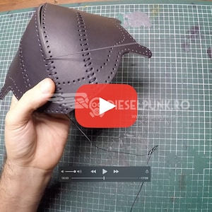 May include: A person is sewing a dark brown leather piece with a needle and thread. The leather piece has holes punched around the edges and is being sewn together. The person is using a green cutting mat as a work surface.