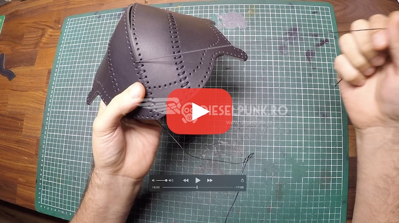 May include: A person is sewing a dark brown leather piece with a needle and thread. The leather piece has holes punched around the edges and is being sewn together. The person is using a green cutting mat as a work surface.