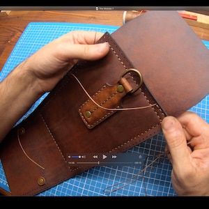 May include: A person is sewing a brown leather bag with a metal ring and a brown leather strap. The bag is on a blue cutting mat.