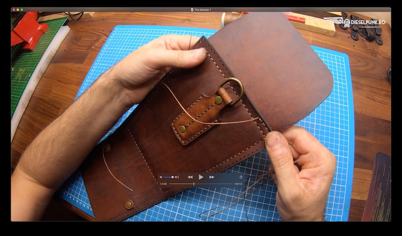 May include: A person is sewing a brown leather bag with a metal ring and a brown leather strap. The bag is on a blue cutting mat.