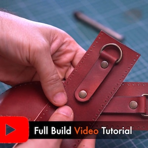May include: Close-up of two pieces of burgundy leather with metal rings and rivets. The leather pieces are being held by a person's hands. The text "Full Build Video Tutorial" is displayed in white on a black background with a red play button.