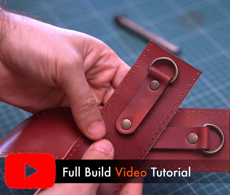 May include: Close-up of two pieces of burgundy leather with metal rings and rivets. The leather pieces are being held by a person's hands. The text "Full Build Video Tutorial" is displayed in white on a black background with a red play button.