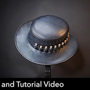 Puede incluir: Un sombrero de cuero azul oscuro con copa plana y ala ancha. El sombrero presenta una banda alrededor de la base decorada con una serie de pequeños frascos de vidrio transparente con tapones de corcho. El texto Pattern and Tutorial Video es visible.