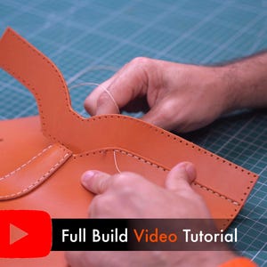 May include: A tan leather piece with pre-punched holes along the edges being hand-stitched with light-colored thread. The leather has a curved flap section and a rectangular body. A red YouTube icon and the text Full Build Video Tutorial appear.