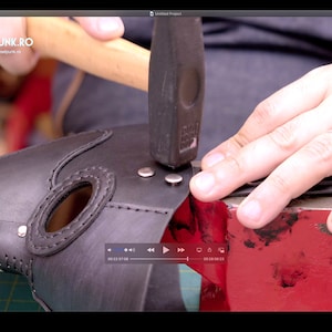 May include: A black leather plague doctor mask is being assembled with rivets. A hammer is used to set the rivets. The mask has an elongated beak shape and is studded with silver rivets around the edges. The mask is on a red anvil.