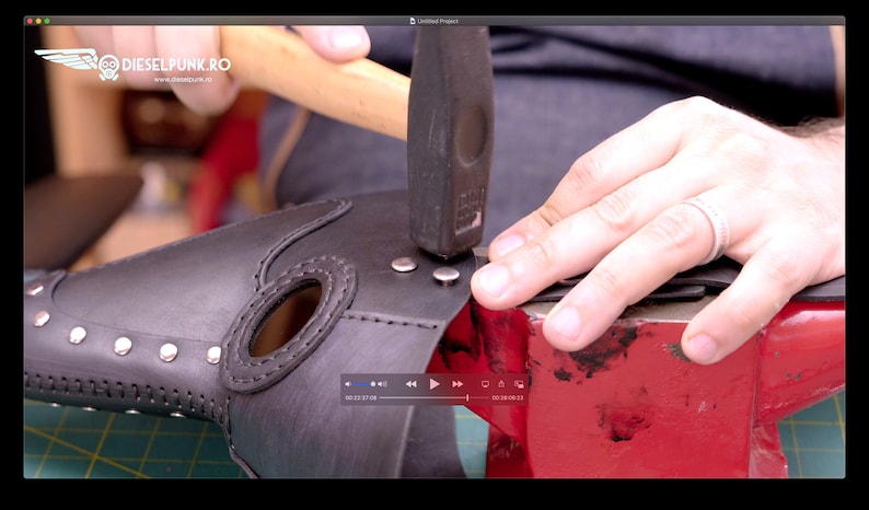 May include: A black leather plague doctor mask is being assembled with rivets. A hammer is used to set the rivets. The mask has an elongated beak shape and is studded with silver rivets around the edges. The mask is on a red anvil.