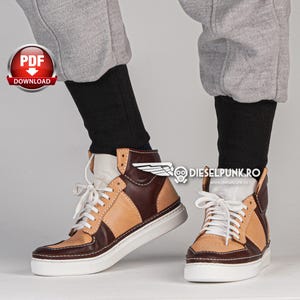 Puede incluir: Un par de zapatillas de ca&ntilde;a alta marrones y beige con cordones blancos. Las zapatillas tienen una suela blanca y una parte superior de cuero marr&oacute;n con un panel de cuero beige en el lateral. Las zapatillas las lleva puestas una persona que lleva pantalones de ch&aacute;ndal grises.