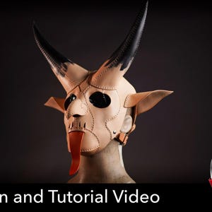 May include: A tan leather mask featuring two large, curved black horns, pointed ears, and a long, protruding red tongue. The mask is constructed with visible stitching along the seams and includes a strap with a buckle for secure fastening.