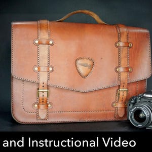 May include: A tan leather satchel with a top handle, two vertical straps featuring brass buckles, and a triangular emblem on the front flap. A black Canon DSLR camera with a lens is positioned to the right of the bag. Text reads Pattern and Instructional Video.