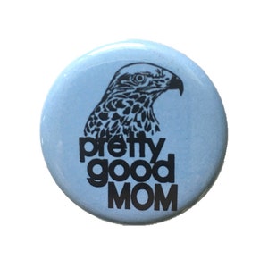 Pode incluir: Um botão azul claro com uma silhueta preta de uma cabeça de falcão e o texto "pretty good MOM" em preto.