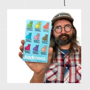 May include: A sticker sheet with nine colorful cat illustrations. Each cat has the text "You can do it, just focus, you're great." The sticker sheet is held by a person wearing a plaid shirt and a hat that says "badknees".