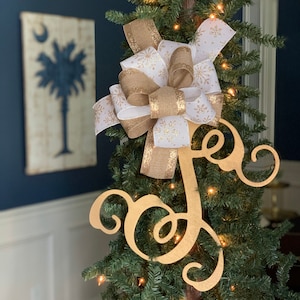 May include: A decorated Christmas tree with a large bow and a gold letter "J" ornament. The bow features white ribbon with gold snowflakes and gold glitter ribbon. The tree is lit with small white lights, creating a festive holiday display.