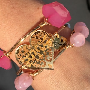 May include: Gold bangle bracelets with pink and light pink beads and a gold charm with a filigree heart design. The charm has the word "stoehr" engraved on it.