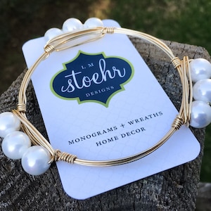Puede incluir: Una pulsera de alambre dorada con detalles de perlas blancas. La pulsera tiene una etiqueta que dice "L.M. Stoehr Designs" y "Monograms + Wreaths Home Decor". La pulsera se muestra sobre una superficie de madera.
