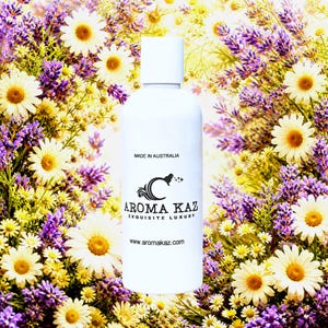 May include: A white cylindrical bottle with a white cap featuring the black text MADE IN AUSTRALIA, the AROMA KAZ logo, the words EXQUISITE LUXURY, and the website www.aromakaz.com, centered against a background of purple lavender and white daisies.