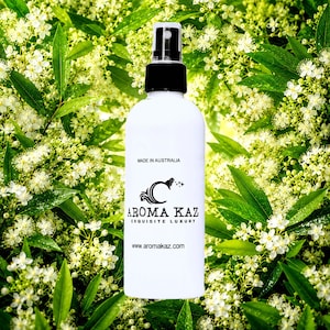 Könnte beinhalten: Weiße Sprühflasche mit schwarzem Deckel, mit dem Logo "Aroma Kaz" und dem Text "Made in Australia". Die Flasche befindet sich vor einem Hintergrund aus grünen Blättern und weißen Blüten. Die Website-Adresse www.aromakaz.com ist ebenfalls sichtbar.