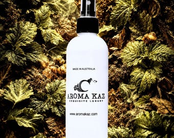 May include: A white spray bottle with a black cap and nozzle, featuring the text "AROMA KAZ EXQUISITE LUXURY" and "MADE IN AUSTRALIA." The bottle is set against a backdrop of green leaves and brown organic matter.
