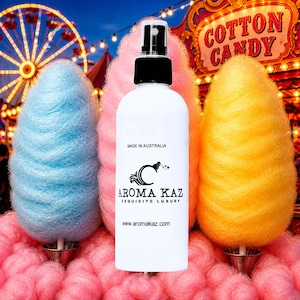 Può includere: Un flacone spray bianco con tappo nero e la scritta "AROMA KAZ EXQUISITE LUXURY" è centrato davanti a zucchero filato blu, rosa e giallo. Lo sfondo presenta una scena di carnevale con una ruota panoramica e un'insegna "COTTON CANDY".