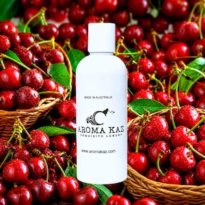 Op de afbeelding: Een witte cilindrische fles met een platte dop, voorzien van de zwarte tekst MADE IN AUSTRALIA, AROMA KAZ, EXQUISITE LUXURY en www.aromakaz.com, geplaatst voor twee gevlochten manden gevuld met talloze verse, glanzende rode kersen.