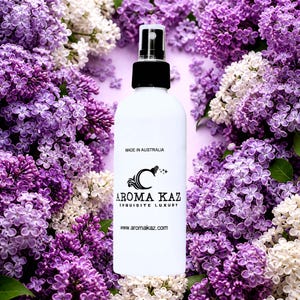 May include: A white spray bottle with a black cap and the text "AROMA KAZ EXQUISITE LUXURY" is surrounded by purple and white lilac flowers. The bottle also has the text "MADE IN AUSTRALIA" and the website address.