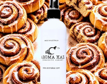 May include: A white spray bottle with a black cap, featuring the text "AROMA KAZ EXQUISITE LUXURY" and "MADE IN AUSTRALIA", is surrounded by freshly baked cinnamon rolls with icing. The website address www.aromakaz.com is also visible.