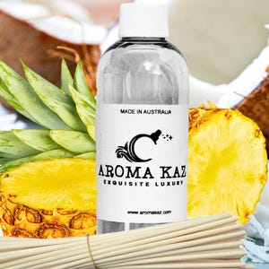Könnte beinhalten: Eine klare Glasflasche mit einem weißen Etikett mit der Aufschrift "AROMA KAZ EXQUISITE LUXURY" und "MADE IN AUSTRALIA". Die Flasche ist von Ananasscheiben, Kokosnuss und Schilfrohrdiffusoren umgeben. Die Website www.aromakaz.com ist ebenfalls sichtbar.