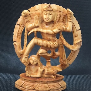 Può includere: Una scultura in legno del dio indù Shiva nella sua forma Nataraja, che danza su un demone. La scultura è scolpita in un colore marrone chiaro e presenta dettagli intricati. La scultura è in piedi su una base con un design decorativo.