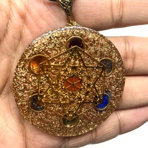 May include: A circular, gold-toned pendant with a Metatron's Cube design, incorporating colourful gemstones. The pendant is suspended from a black cord. The pendant is held in a hand.