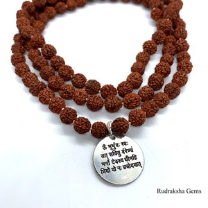 May include: A brown rudraksha mala or japa mala prayer beads necklace with a silver pendant featuring a Sanskrit mantra. The mantra is "Om Bhurbhuvah Svah Tat Savitur Varenyam Bhargo Devasya Dhimahi Dhiyo Yo Nah Prachodayat".