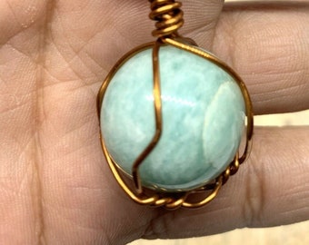 May include: A light blue gemstone pendant wrapped in copper wire, hanging from a black cord necklace. The pendant is a round, smooth stone with a subtle shimmer. The necklace is a simple, elegant design.