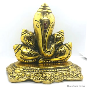 Puede incluir: Una estatua dorada de la deidad hind&uacute; Ganesha, con una cabeza de elefante y m&uacute;ltiples brazos. La estatua tiene detalles intrincados y est&aacute; colocada sobre una base decorativa. Tres gemas rojas est&aacute;n engastadas en la frente.