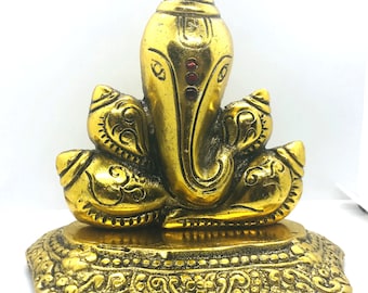May include: A gold-colored statue of the Hindu deity Ganesha, with a large elephant head and multiple arms. The statue has intricate details and is set on a decorative base. Three red gems are set in the forehead.