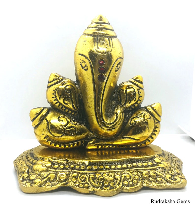 Puede incluir: Una estatua dorada de la deidad hind&uacute; Ganesha, con una cabeza de elefante y m&uacute;ltiples brazos. La estatua tiene detalles intrincados y est&aacute; colocada sobre una base decorativa. Tres gemas rojas est&aacute;n engastadas en la frente.