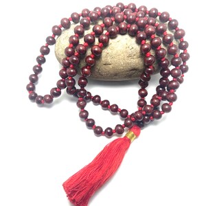 May include: A red wooden mala bead necklace with a red tassel. The beads are strung on a red cord and the tassel is attached to the end of the cord.