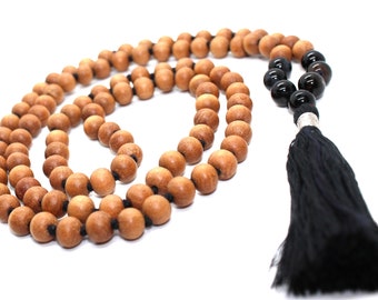 Puede incluir: Un collar de cuentas mala de madera con un borla negro y cuentas de ónix negro. El mala está hecho de cuentas de sándalo y tiene un cordón negro.
