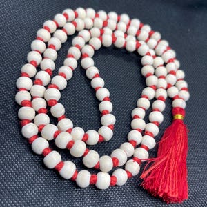 May include: A strand of round, off-white beads strung on a vibrant red cord. The beads are arranged in a circular pattern, and the necklace features a prominent red tassel at one end, secured by a small, shiny gold-coloured metal band.
