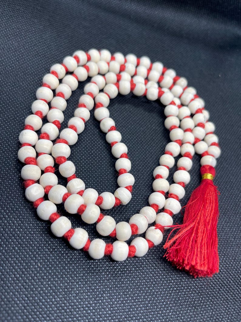 May include: A strand of round, off-white beads strung on a vibrant red cord. The beads are arranged in a circular pattern, and the necklace features a prominent red tassel at one end, secured by a small, shiny gold-coloured metal band.