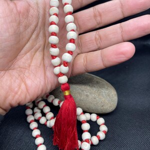 May include: A string of prayer beads featuring smooth, off-white spherical beads separated by knots on a vibrant red cord. The strand terminates in a prominent, deep red tassel accented with a small, metallic gold band at the top of the fringe.