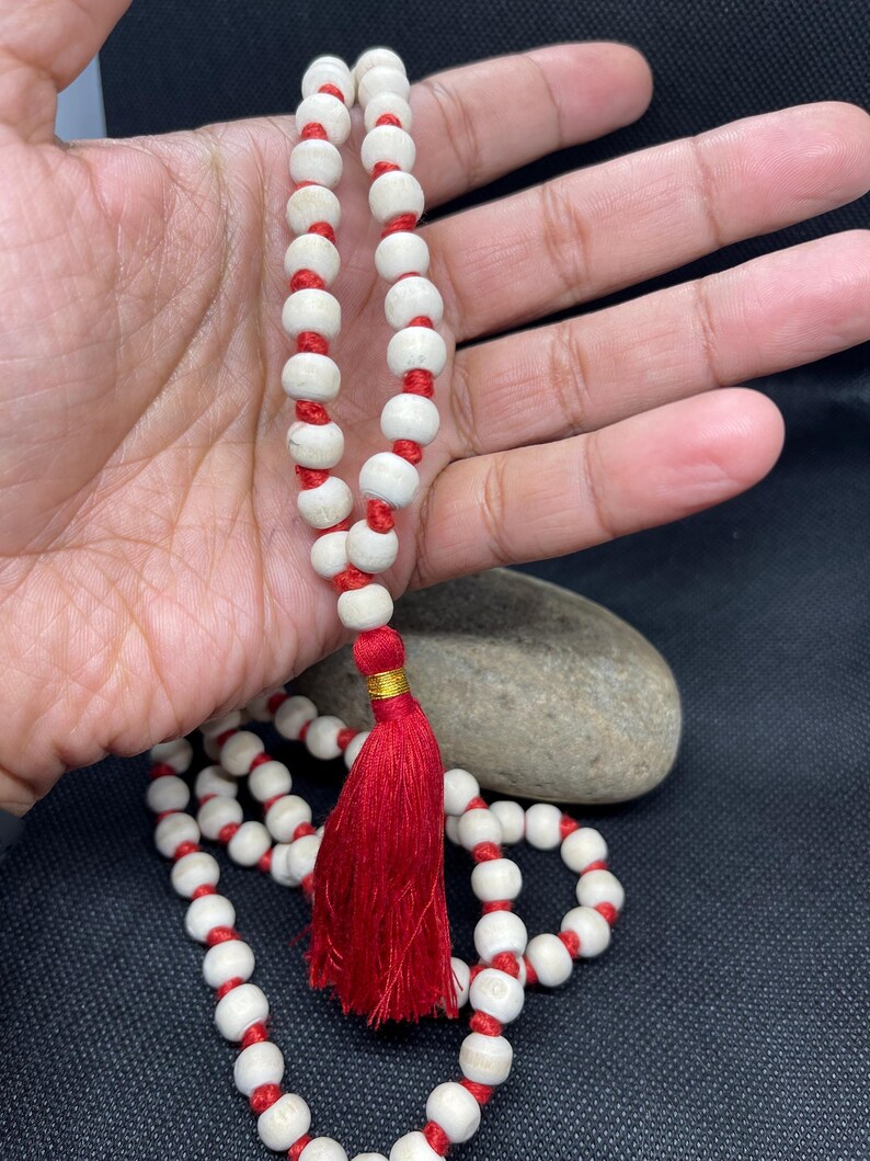 May include: A string of prayer beads featuring smooth, off-white spherical beads separated by knots on a vibrant red cord. The strand terminates in a prominent, deep red tassel accented with a small, metallic gold band at the top of the fringe.