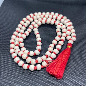 May include: A strand of round, off-white beads strung on a vibrant red cord, arranged in a circular loop. The necklace features a prominent red tassel at the bottom, secured with a small gold-coloured band, creating a continuous, knotted design.